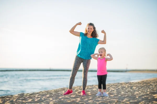 Happy mother and daughter in fitness gear on beach flexing arms — Stockfoto