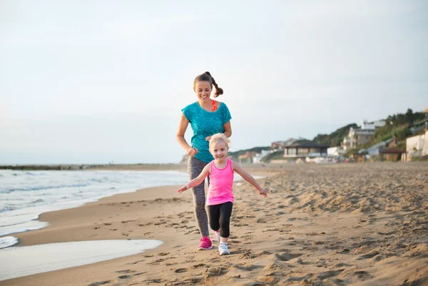 Fit, glad mor kører bag ung datter på stranden - Stock-foto
