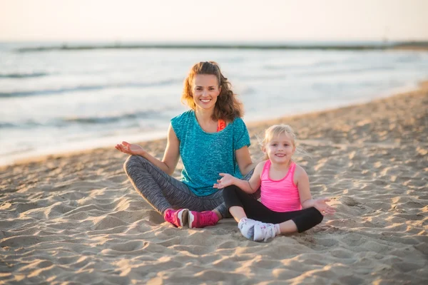 Happy mother and daughter sitting in lotus position on beach — Stock Photo, Image
