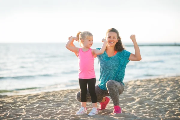 Young mother and daughter in fitness gear on beach flexing arms — Zdjęcie stockowe