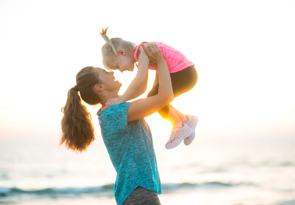 Lykkelig mor holder barn op i luften på stranden ved solnedgang - Stock-foto