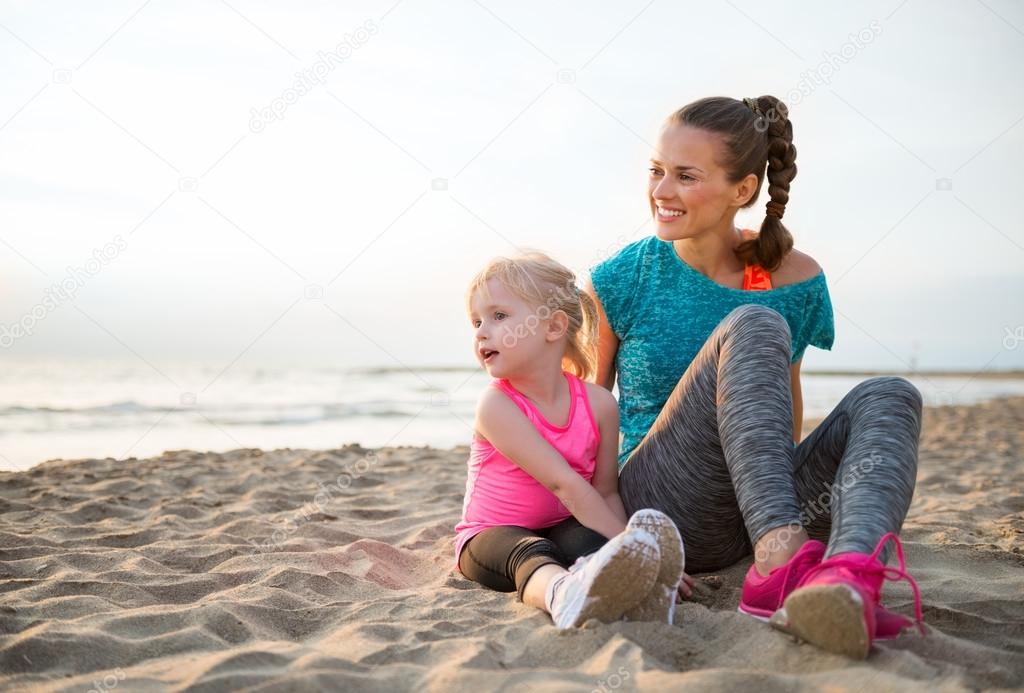 Smiling, fit mother sitting next to young daughter on the sand