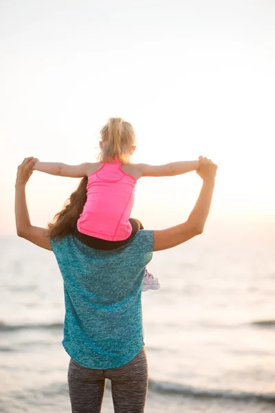 Mother holding young daughter on shoulders at sunset on beach — Stock Photo, Image