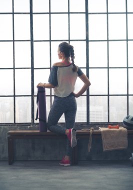 Rear view of fit woman with yoga mat looking out loft gym window
