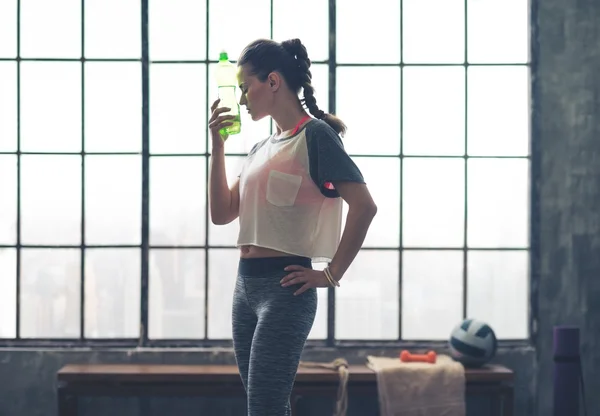 Woman by window in loft gym holding water bottle to head — 스톡 사진