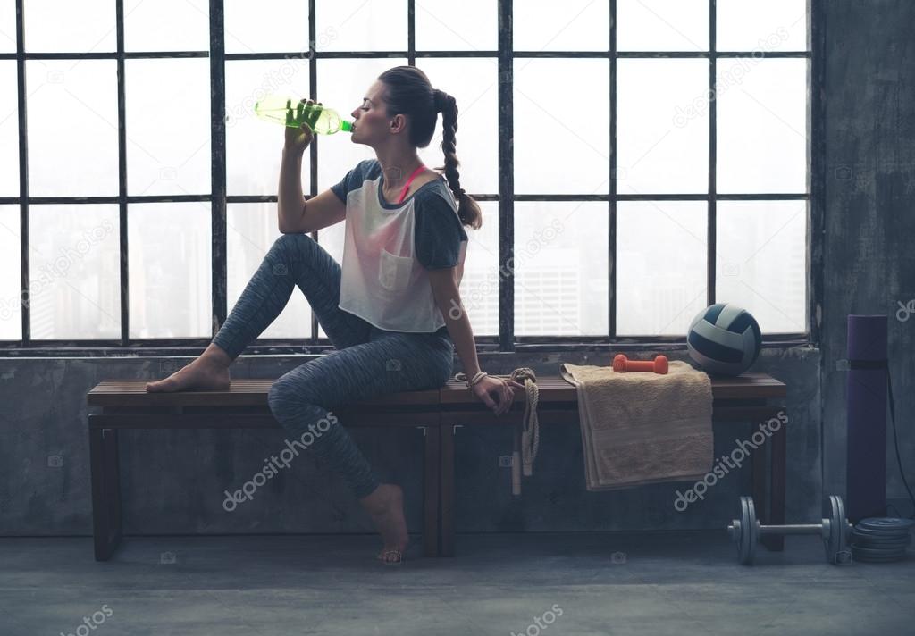 Fit woman in profile sitting on bench in loft gym drinking water