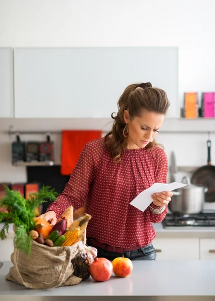 Mulher na cozinha comparando lista de compras com itens no saco — Fotografia de Stock