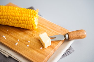 Corn cob on wooden board with pat of butter on corn skewer