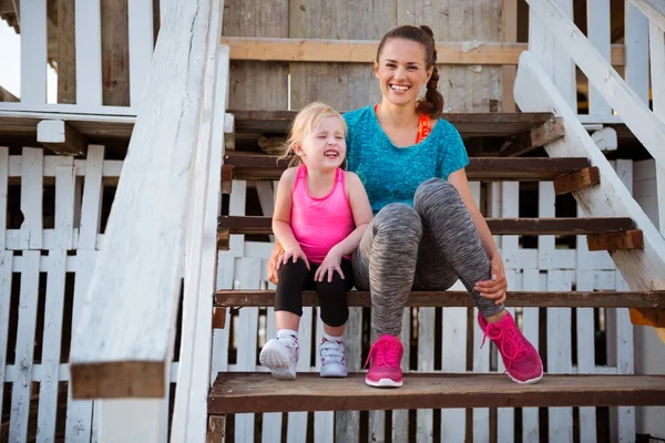 Mother and daughter in fitness gear sitting on beach hut steps — Stock Photo, Image