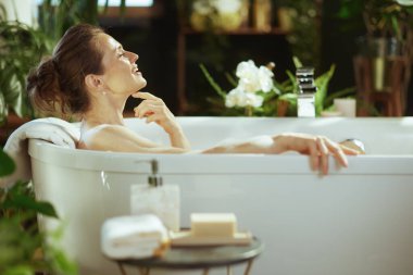 Orta yaşlı bir kadın, saçı topuz içinde, beyaz köpük banyosunda huzur buluyor, gülümsüyor, gözleri kapalı, eli çenesinde. Yeşil bitkiler ve yumuşak ışık, sakin, spa benzeri bir kişisel bakım atmosferi oluşturur.