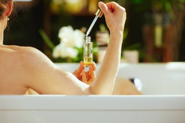 Arkadan görülen bir kadın, beyaz bir küvete damlatarak sarı banyo sıvısı dağıtıyor. Serene bulanık yeşil yapraklar ve beyaz çiçekler sükunet ve kişisel bakım havasını artırır.