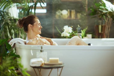 Çörekleri olan orta yaşlı bir kadın gözleri kapalı, mutlu bir gülümsemeyle lüks bir köpük banyosunda rahatlıyor. Etrafı yemyeşil bitkilerle çevrili olan bu bitki, özgüvene ve refaha odaklı sakin bir ev spa 'sını çağrıştırıyor..