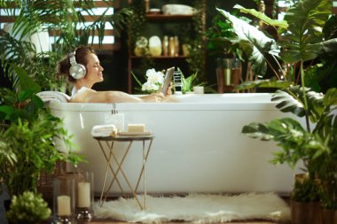 Orta yaşlı bir kadın, yemyeşil, bitki dolu bir küvette, gözleri kapalı, kulaklık takmış ve elinde tablet tutan huzurlu bir yalnızlık bulur. Bu sakin spa benzeri banyo rahatlama ve kişisel bakım için idealdir..