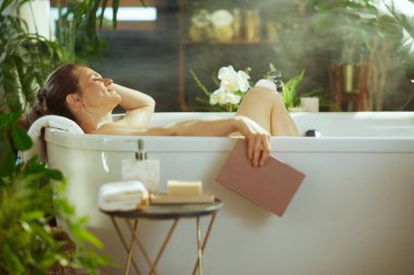 Kapalı gözlü sakin bir kadın ve lüks beyaz bir küvette gevşeyen nazik bir gülümseme. Etrafı yemyeşil bitkilerle, buharla ve doğal ışıkla çevrili bir şekilde, elinde bir kitapla huzurlu bir kişisel bakım anından zevk alıyor.