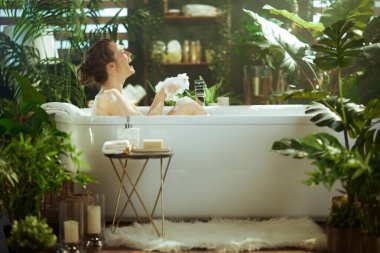 Çörekli mutlu bir kadın, modern beyaz köpük banyosunda yüksek sesle gülüyor, neşe içinde köpük tutuyor, dingin ve lüks bir spa ortamında yemyeşil bitkilerle çevrili..