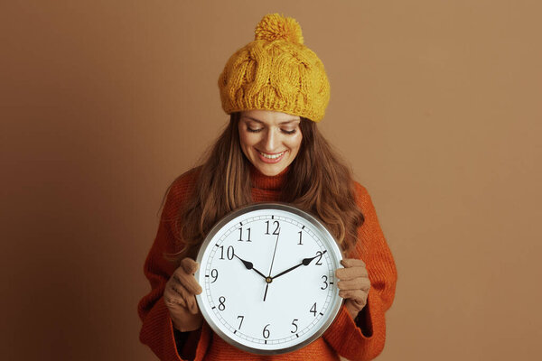 A smiling woman in a yellow beanie and orange turtleneck sweater holds a large wall clock, looking down at it. Her warm, cheerful expression against a brown background evokes a cozy autumn mood.