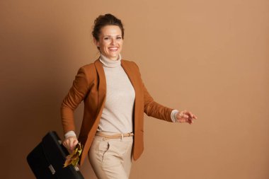 Ceketli ve balıkçı yakalı neşeli bir iş kadını elinde siyah bir evrak çantası ve renkli sonbahar yapraklarıyla güvenle yürüyor. Doğrudan gülümsüyor, profesyonel başarıyı ve moda düşüşünü simgeliyor.