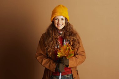 Sarı bereli, kahverengi ceketli ve kırmızı kazaklı gülümseyen bir kadın, canlı bir buket yaprak tutuyor. Görüntü neşeli ve sıcak bir sonbahar havası yakalıyor, mevsimlik temalar için mükemmel..