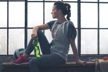 Woman in workout gear sitting on bench in city loft looking out clipart