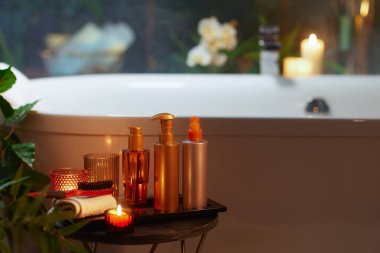 Bir banyo ve vücut ürünleri koleksiyonu, bir fırça ve bir havlu koyu bir tepsi ve masa üzerinde güzelce düzenlenir. Bulanık mumlar ve yeşil yeşilliklerden gelen yumuşak, sıcak ortam ışığı ön planı aydınlatarak huzur verici, eve kaplıca deneyimi yaratıyor..