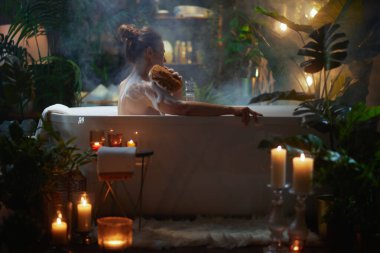 Yeşil bitkiler ve mum ışığıyla çevrili banyo köpüğü kullanan bir kadın. Lüks bir kaplıcada rahatlatıcı bir kişisel bakım ritüeli.