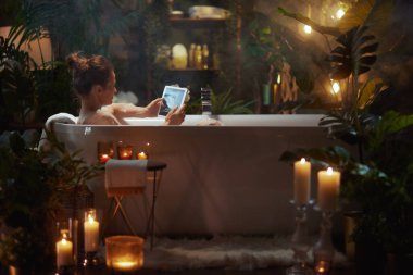 Köpük banyosunda elinde tablet tutan bir kadın. Etrafı mumlar ve yeşilliklerle çevrili. Rahatlamayı, lüksü ve teknolojik bir çarpıtmayla modern kendine bakımı çağrıştırıyor..