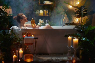 Bir kadın köpük banyosunda, elinde altın bir şişeyle bitki ve mum ışığında rahatlıyor, aromaterapinin tadını çıkarıyor ve huzurlu bir spa deneyimi yaratıyor..
