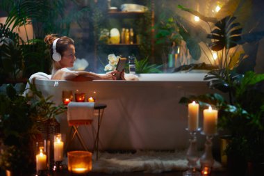 Köpük banyosu yapan, kulaklık takan ve tablet kullanan, bitkiler ve mumlarla çevrili, kendine bakım ve sükuneti somutlaştıran bir kadın..