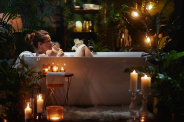Bir kadın, mumlar ve bitkilerle çevrili köpük banyosunda dekoratif bir içecekten hoşlanır. Ilık ışıklandırma, rahatlama, kendine bakım ve hoşgörüyü çağrıştırıyor..
