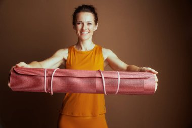 Hardal rengi kolsuz tişörtlü ve uyumlu taytlı gülümseyen bir kadın pembe yoga minderini yatay olarak tutuyor, kolları açık. Kahverengi bir arka planda refah ve aktivite sunar..