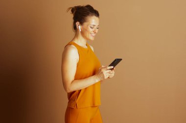 Hardal sarısı atletik giysili ve kulaklıklı gülümseyen orta yaşlı bir kadın profilden akıllı telefonuna dikkatle bakarken görülüyor. Dijital bağlılık ve aktif, modern yaşam tarzını somutlaştırıyor..