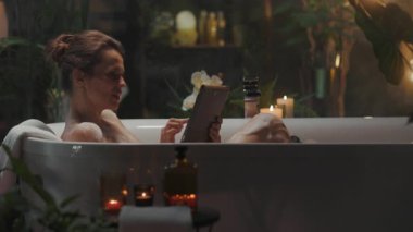Orta yaşlı bir kadın, loş ve rahat bir banyoda elinde tabletle huzurlu bir banyonun tadını çıkarıyor. Mumlar, bitkiler ve kehribar şişelerle süslenmiş sahne, konfor ve kendine bakımı somutlaştırıyor..
