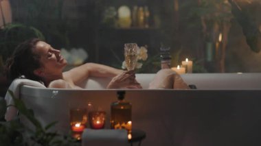 Saçları topuz şeklinde olan bir kadın lüks ve sofistike bir kişisel bakım anından zevk alır, köpüklü bir küvette şampanya yudumlarken. Mumlar ve bitkilerle süslenmiş loş bir banyo huzurlu ve davetkar bir akşam ritüeli yaratıyor..