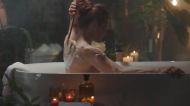 Arkadan bakıldığında, kumral saçlı bir kadın, küvette uzun saplı bir fırçayla omzunu fırçalayarak rahatlatıcı bir kişisel bakım ritüelinden zevk alır. Mumlar ve yemyeşil çiçeklerle çevrili, sükuneti kucaklıyor..