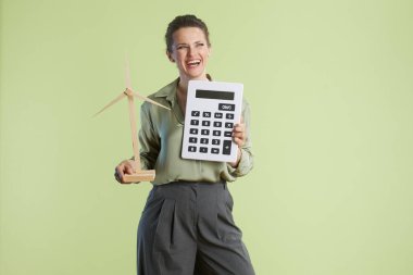 Bir kadın oyuncak bir hesap makinesi ve rüzgar türbini modeli tutuyor. Bu da yenilenebilir enerji, finansal planlama ve pozitif bir çevresel görünüm öneriyor..