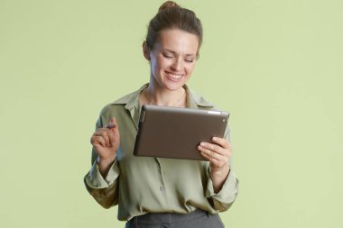 Gülümseyen orta yaşlı bir kadın, yeşil bir zemine dayanan yakalı gömlek ve gri pantolon giyerek tabletin içeriğinden zevk alır..