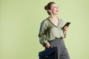 Akıllı telefonu ve çantası olan gülümseyen bir kadın, neşeli bir ifadeyle yukarıya bakıyor. Açık yeşil bluz ve gri pantolon yumuşak yeşil zemine karşı başarıyı işaret ediyor..