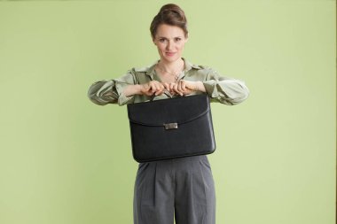 Bir kadın bir çanta tutuyor, kendine güveni tam. Yeşil bluz ve gri pantolonla, yeterliliğini ve kariyer başarısını temsil ediyor..