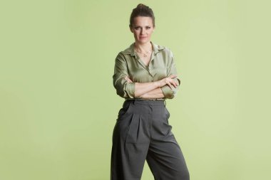 Kollarını kavuşturmuş, yeşil saten bluz ve gri pantolon giyen orta yaşlı bir kadın açık yeşil bir zeminde güven ve profesyonellik yayıyor..