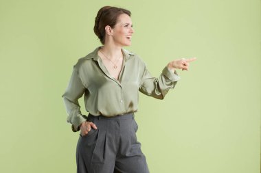 İpeksi yeşil gömlekli ve gri pileli pantolonlu gülümseyen bir kadın sağı işaret ederek yumuşak yeşil bir arkaplana karşı güven ve ulaşılabilirlik taşıyor. İletişim kavramları için ideal.