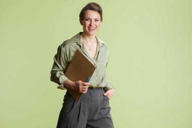 30 'lu yaşlarda gülümseyen bir kadın bir elinde kağıt ve kalem, bir elinde de cebinde. Yeşil bir gömlek ve gri bir pantolon giyiyor, açık yeşil bir arka plana karşı..