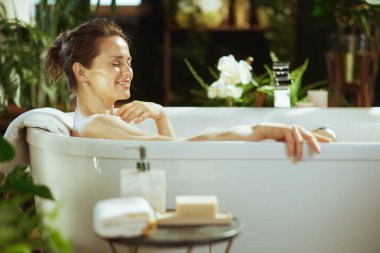 Orta yaşlı, şık bir kadın beyaz köpük banyosunda mutluluğu buluyor, gülümsüyor, gözleri kapalı. Yeşilli yeşil bitkiler ve beyaz orkideler, lüks rahatlığı ön plana çıkaran sakin bir zemin oluştururlar.