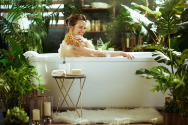 Beyaz bir küvette gülümseyen bir kadın köpüklerin arasında omzunu doğal bir süngerle ovalıyor. Yeşil bitkilerle süslenmiş banyo ve mum ışığı huzurlu, lüks bir spa atmosferi yaratıyor..