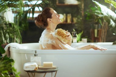 Gözleri kapalı ve huzurlu bir gülümsemesi olan bir kadın, omuzlarını doğal bir süngerle yıkayarak köpük banyosundan zevk alır. Bitki dolu banyo dinlenmek için sakin, spa gibi bir sığınak yaratıyor..