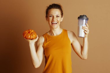 Hardal sarısı bluzlu gülümseyen bir kadın, düşmekte olan bir balkabağı ve protein karıştırıcısı, somut sağlık, sağlık ve sonbahar sezonunu elinde tutar..