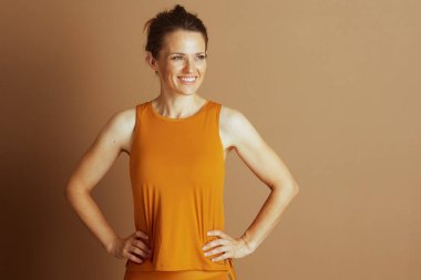 Kahverengi saçlı bir kadın, hardal sarısı atletik bir bluz ve alt giyen, elleri kalçalarında, tarafsız kahverengi bir stüdyo arka planına karşı gülümseyen, vücut bulmuş ve kendine güveni olan.