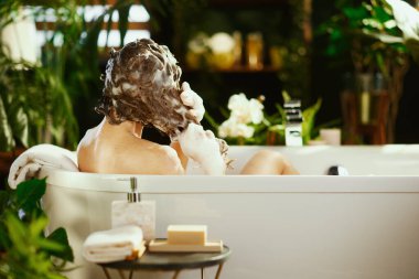 Kişi saçlarını yemyeşil bitkilerle çevrili beyaz bir küvette şampuanla yıkar. Yakınlardaki beyaz havlular ve banyo ürünleri kişisel bakım ve rahatlama için sakin, spa benzeri bir atmosfer yaratıyor.