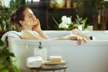 Orta yaşlı bir kadın, gözleri kapalı ve sakin, beyaz bir köpük banyosunda huzur buluyor, elleri yanağına dokunuyor. Bereketli yeşil bitkiler, beyaz orkideler ve yumuşak ışıklandırma kendi kendine bakım için lüks bir kaplıca ortamı oluşturur