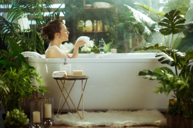 Profilde bir kadın, sakin bir ifadeyle gözleri kapalı, köpük banyosuna dalmış, köpük tutmuş, bitkilerle dolu yumuşak, sisli, kendine özen gösteren bir banyoda..