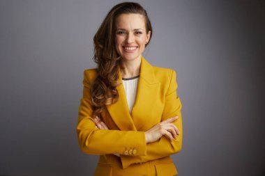 Sarı ceketli kendine güvenen bir iş kadını kollarını kavuşturmuş bir şekilde gülümseyerek profesyonel bir stüdyo fotoğrafında doğrudan kameraya bakar..